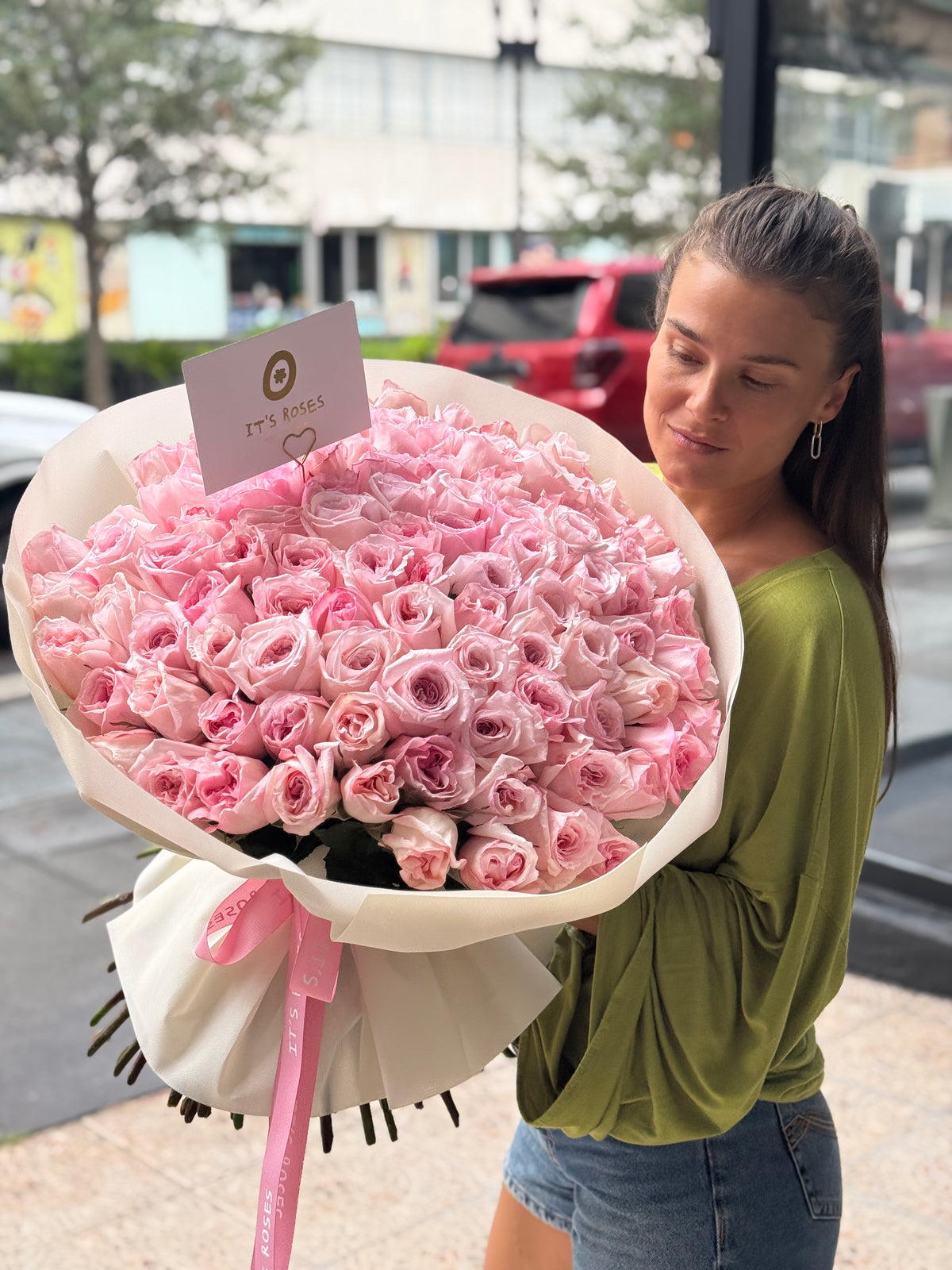 A woman holds a large bouquet of pink roses wrapped in white paper with a pink ribbon and a small card that reads 'IT'S ROSES'. 